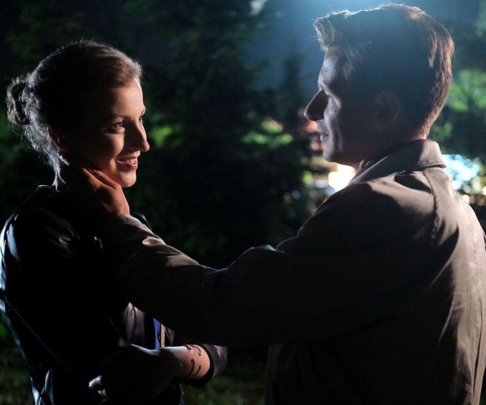 Natalka (Dominika Suchecka) i Adam Karski (Patryk Szwichtenberg) z serialu „M jak miłość” patrzą na siebie z uśmiechem, Adam obejmuje jej szyję, tworząc romantyczną scenę w nocnym, zielonym otoczeniu. Więcej o ich losach na Super Seriale.