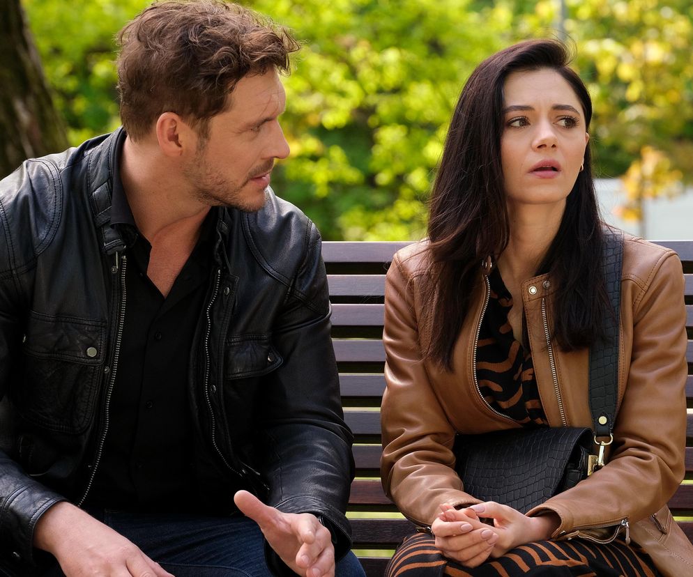 Aktorzy Mikołaj Roznerski jako Marcin i Adriana Kalska jako Iza siedzą na ławce. Marcin w czarnej kurtce z zainteresowaniem patrzy na Izę w brązowej kurtce, która ma zmartwioną minę. Scena z serialu M jak miłość. Więcej o tym na Super Seriale.