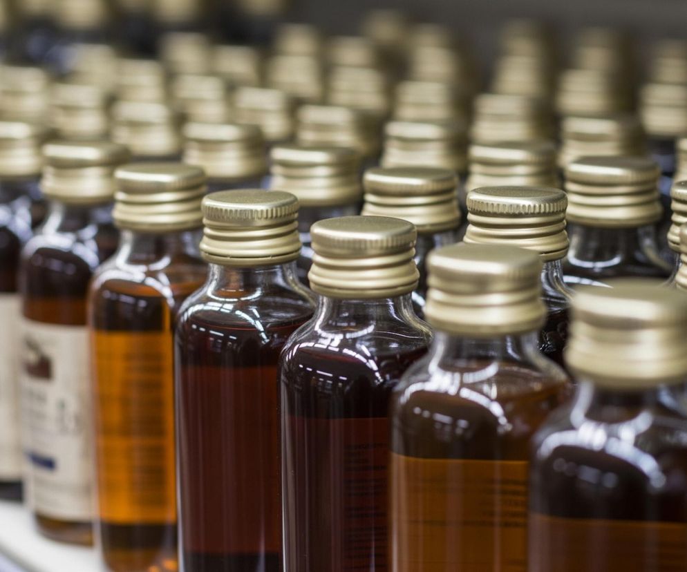 Rząd szklanych butelek z ciemną zawartością, każda z metalową, złotą nakrętką, stoi na białej półce. Bliżej aparatu znajdują się butelki wypełnione cieczą o bursztynowym lub ciemnobrązowym odcieniu, wyraźnie widoczne są również etykiety na niektórych z nich, choć ich treść jest nieczytelna. W tle widać kolejne rzędy identycznych butelek, które stopniowo stają się rozmyte i mniej szczegółowe. Oświetlenie z góry odbija się na powierzchniach nakrętek i ściankach butelek, tworząc jasne refleksy.