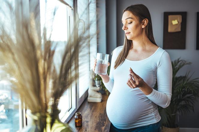 Kobieta w ciąży stojąca przy oknie z tabletkami w dłoni