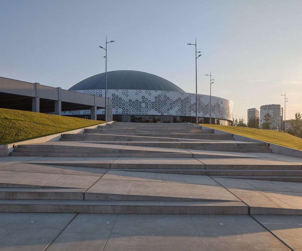 Nowoczesna hala widowiskowo-sportowa Urania w Olsztynie, autorstwa URBAN ARCHITECT, z odnowioną kopułą i szklaną fasadą z geometrycznymi panelami, widoczna od strony rozłożystych schodów. Więcej o projekcie przeczytasz na Architektura Murator Plus.