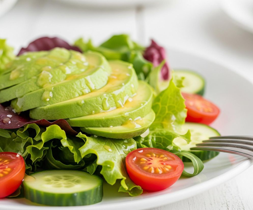 Na pierwszym planie, z lewej strony, widoczny jest talerz wypełniony świeżą sałatką. Dominują liście zielonej sałaty, uzupełnione ciemnoczerwonymi liśćmi, a na wierzchu ułożone są plastry awokado polane klarownym dressingiem z widocznymi nasionkami sezamu. Po bokach sałatki rozmieszczone są plasterki ogórka i połówki pomidorów koktajlowych, a z prawej strony częściowo widoczny jest metalowy widelec. Całość spoczywa na białym, drewnianym blacie o wyraźnej fakturze desek.