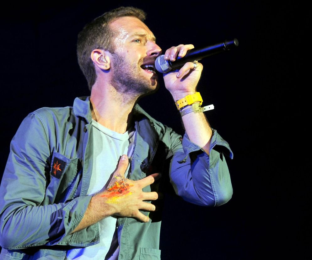 Chris Martin śpiewa do mikrofonu i trzyma rękę na sercu. O micie rockmana przeczytasz na EskaRock.