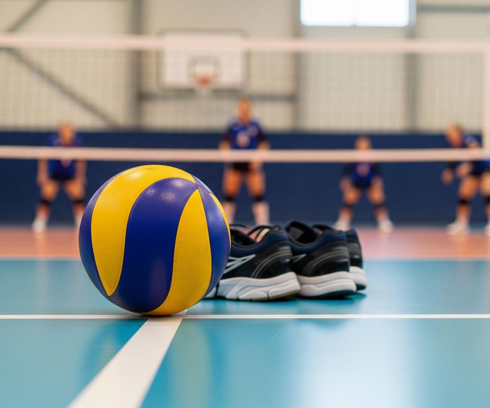 Na pierwszym planie, centralnie, leży piłka do siatkówki o żółto-niebieskich pasach, ustawiona na białej linii wyznaczającej pole gry na jasnoniebieskim parkiecie. Za piłką, po prawej stronie, widać trzy pary ciemnych sportowych butów z białymi podeszwami. W tle rozciąga się boisko do siatkówki z rozciągniętą białą siatką, a za nią, niewyraźnie, widać cztery osoby w niebieskich koszulkach, prawdopodobnie zawodniczki, oraz fragmenty hali sportowej z widocznym koszem do koszykówki.