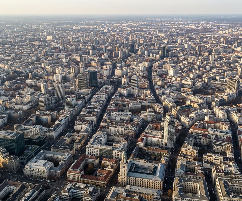 Panoramiczne ujęcie z lotu ptaka przedstawia rozległą metropolię rozciągającą się aż po horyzont. Dominują liczne budynki, głównie w odcieniach szarości, bieli i beżu, z widocznymi, często płaskimi lub lekko spadzistymi dachami. Wśród zwartej zabudowy dostrzegalne są ciemne pasma dróg i pojedyncze, ciemne, wysokie budynki, wyróżniające się na tle jaśniejszej reszty. Horyzont u góry obrazu jest jasny, przechodzący w mlecznobiałe niebo, co sugeruje wczesną porę dnia lub dużą odległość.