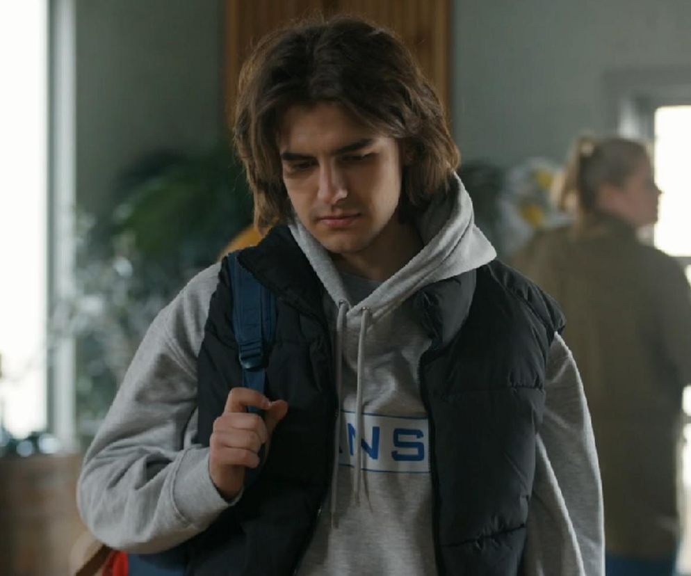 Młody Romeo (Jacek Tyszkiewicz) w szarej bluzie z kapturem i czarnej kamizelce, z plecakiem na ramieniu. Wyraz twarzy aktora wskazuje na zamyślenie i smutek. W tle widać postać Aldony (Elżbieta Romanowska). Więcej o odcinku 3242 serialu Barwy szczęścia przeczytasz na Super Seriale.