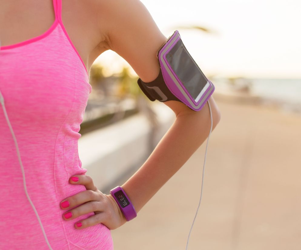 Kobieta w sportowym stroju z opaską na ramię na smartfon i fioletową opaską fitness na nadgarstku, symbolizująca aktywność fizyczną z aplikacją Endomondo. Dowiedz się, jak działa Endomondo i poznaj jego funkcje na Poradnik Zdrowie.