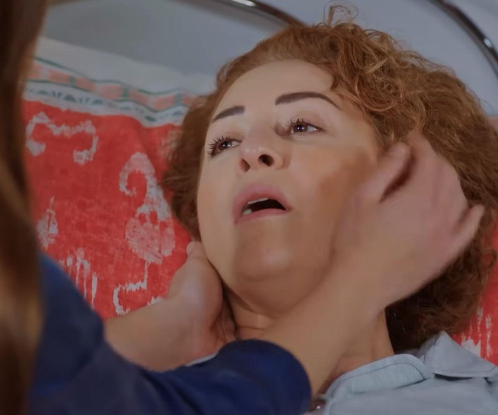 Kobieta z rudymi, kręconymi włosami leży na łóżku w szpitalu, a jej szyję i skroń delikatnie dotyka czyjaś dłoń, wyrażając troskę i współczucie. W tle widoczny jest wzorzysty, czerwony materiał, prawdopodobnie koc. Więcej o losach bohaterów przeczytasz na Super Seriale.