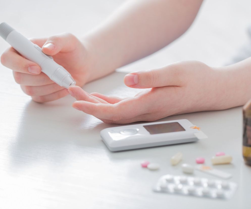 Dłoń osoby mierzącej poziom cukru glukometrem, na białym tle, obok leżą tabletki i butelki z lekami. Mierzenie cukru ma stać się łatwiejsze dzięki nowemu lekowi przezskórnemu, o czym przeczytasz na portalu Poradnik Zdrowie.