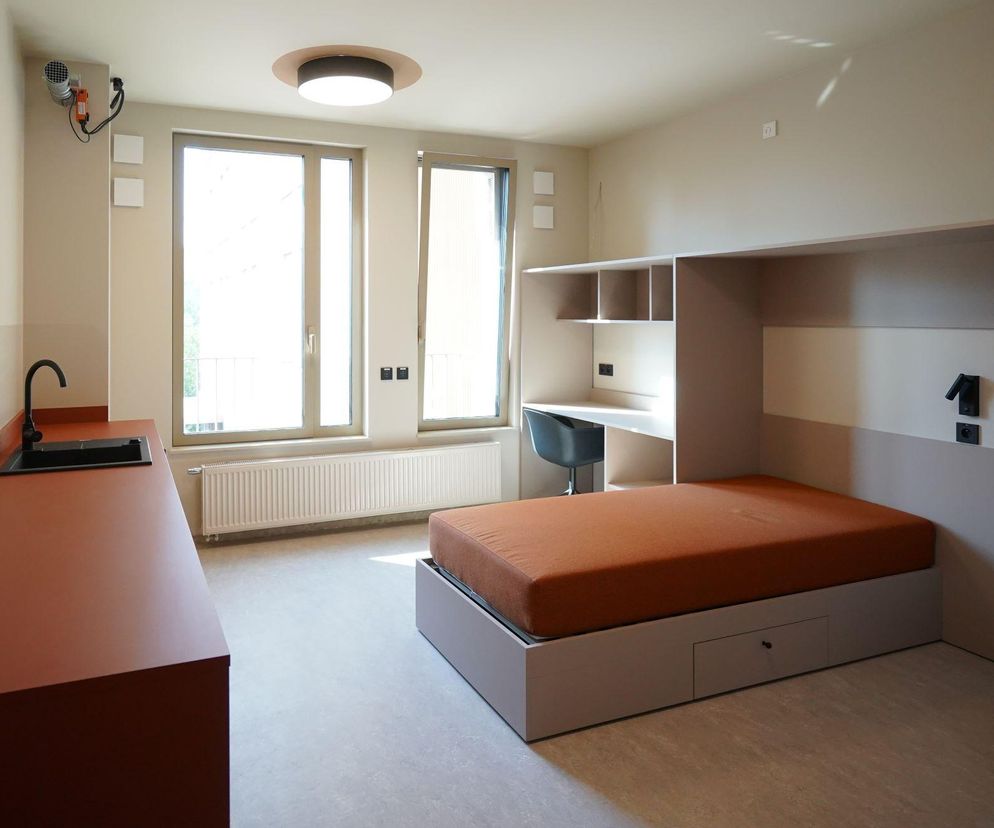 Nowoczesny pokój w akademiku Sulima z brązowym blatem, zlewem, łóżkiem, biurkiem i szafką. To idealne miejsce do studiowania, o cenach którego możesz przeczytać na Super Biznes.