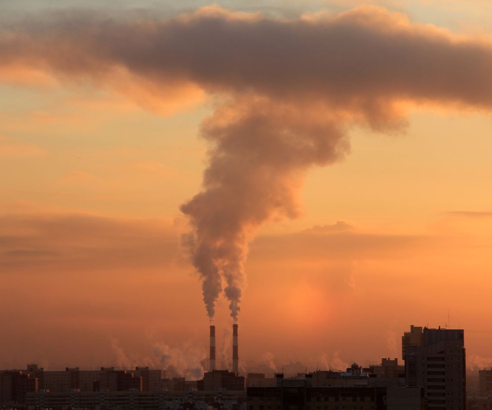 Smug dymu i par buchający z dwóch wysokich kominów, górujących nad miastem o zachodzie słońca. Niebo w odcieniach pomarańczy i różu z chmurami, które częściowo zakrywają słońce. W tle widać zarysy budynków mieszkalnych. Na Poradnik Zdrowie znajdziesz informacje o smogu.