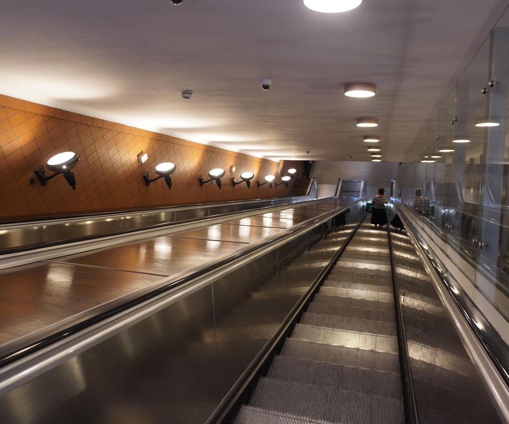 Szerokie schody ruchome w tunelu stacji metra z brązowymi panelami ściennymi i oświetleniem punktowym. Jedna osoba z torbą jedzie w górę, co symbolizuje codzienny ruch miejski. Więcej o infrastrukturze miejskiej przeczytasz na portalu Architektura Murator Plus.
