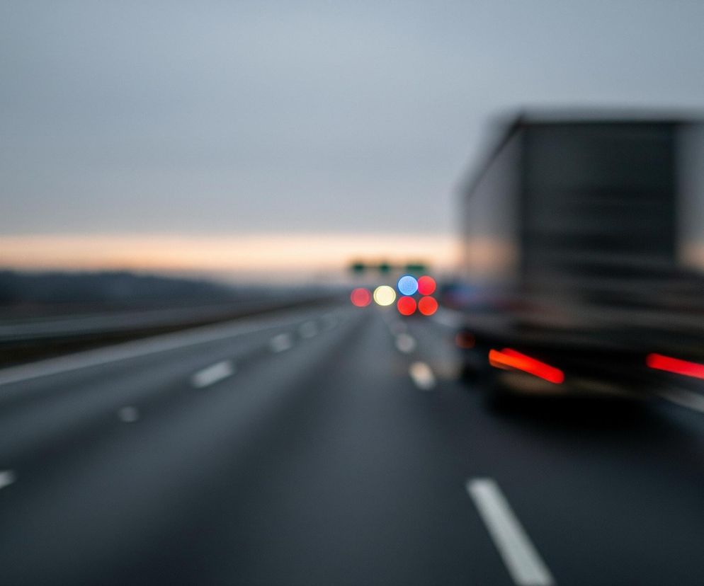 Autostrada z trzema pasami ruchu, po której poruszają się pojazdy, jest widoczna na pierwszym planie i w centrum obrazu. Po prawej stronie autostrady jedzie ciężarówka z włączonymi czerwonymi światłami pozycyjnymi, a w tle widać inne pojazdy z rozmazanymi, okrągłymi światłami w kolorach czerwonym, żółtym i niebieskim. Szare, zachmurzone niebo rozciąga się nad horyzontem, gdzie ciemny las graniczy z jasnym pasem pomarańczowo-różowego światła.