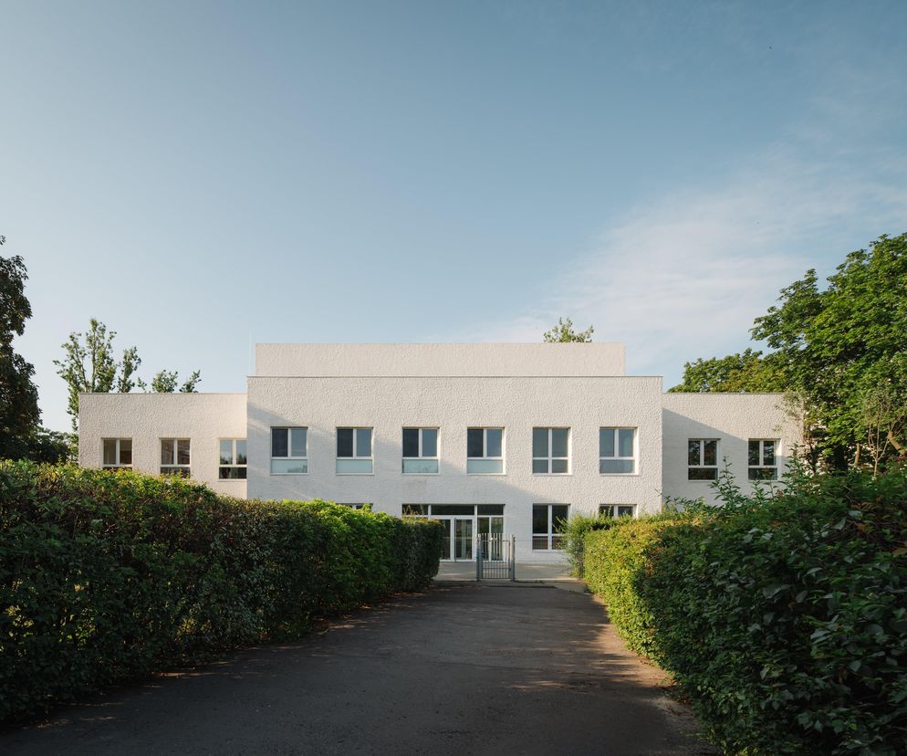 Biały, modernistyczny budynek Żłobka 31 w Warszawie, projektu Asman Pieniężny Architekci sp. z o.o., otoczony zielonymi żywopłotami. Centralne wejście z przeszklonymi drzwiami, nad którym widnieją prostokątne okna. Powierzchnie fasady mają chropowatą teksturę, co dodaje im charakteru. Architektura Murator Plus poleca ten obiekt.