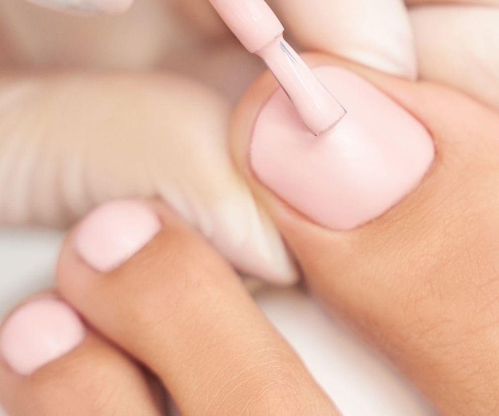 Pedikiur z malowaniem paznokci na pastelowy róż, wykonywany przez osobę w białych rękawiczkach. Detal zabiegu, który wymaga odpowiednich przygotowań, o czym przeczytasz na Poradnik Zdrowie. Unikaj golenia nóg tuż przed.