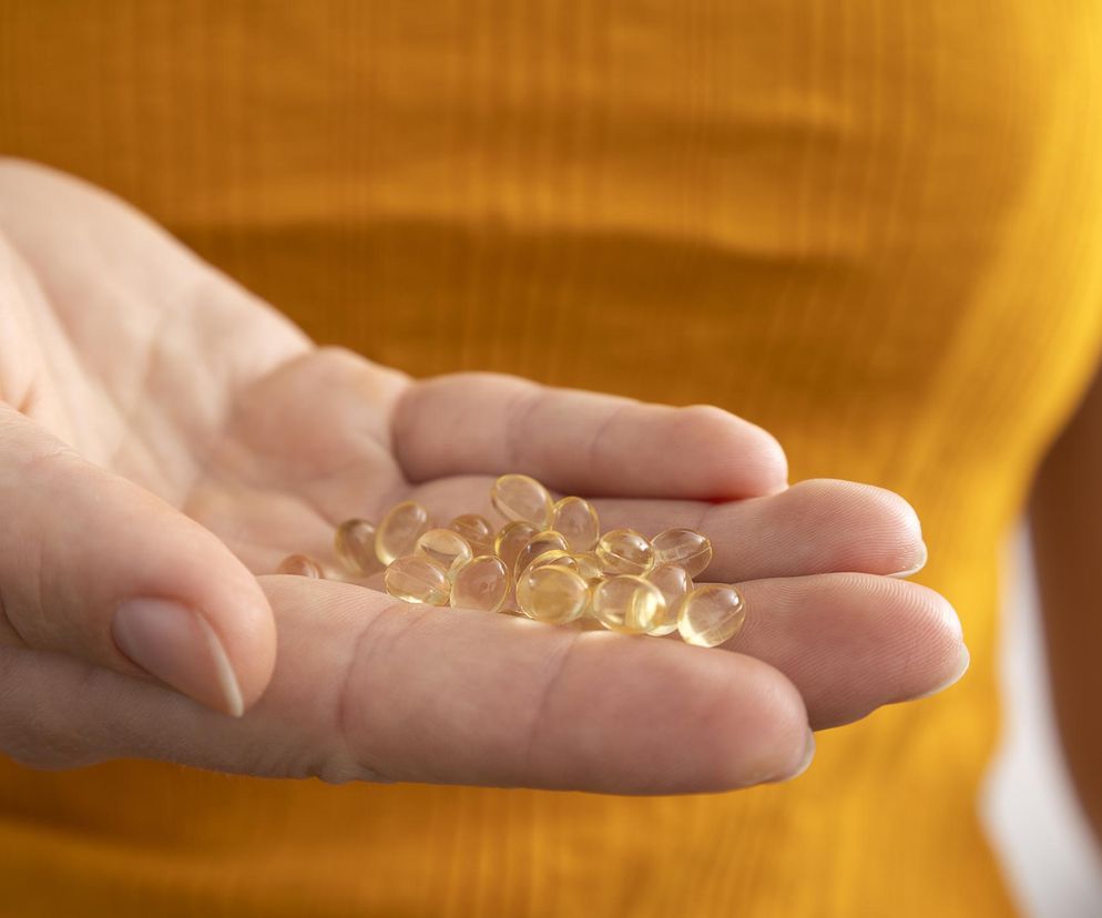 Dłoń kobiety w żółtej bluzce trzyma garść jasnożółtych kapsułek z witaminą D, symbolizujących suplementację. Ostrzeżenia farmaceutów dotyczące łączenia witaminy D z niektórymi lekami, przeczytasz na Poradnik Zdrowie.