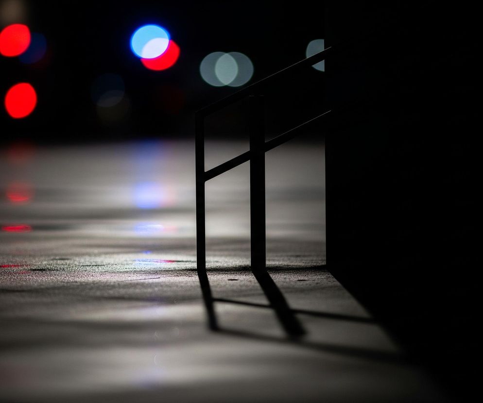 Prawa strona obrazu jest całkowicie ciemna, podczas gdy lewa i górna część przedstawiają rozmyte światła w kolorach czerwonym, niebieskim i białym. Na pierwszym planie, w środkowej części, widać czarną, metalową barierkę lub poręcz, która rzuca długi, ostry cień w prawo. Powierzchnia pod barierką jest ciemna i lekko połyskująca, z widocznymi refleksami świateł.