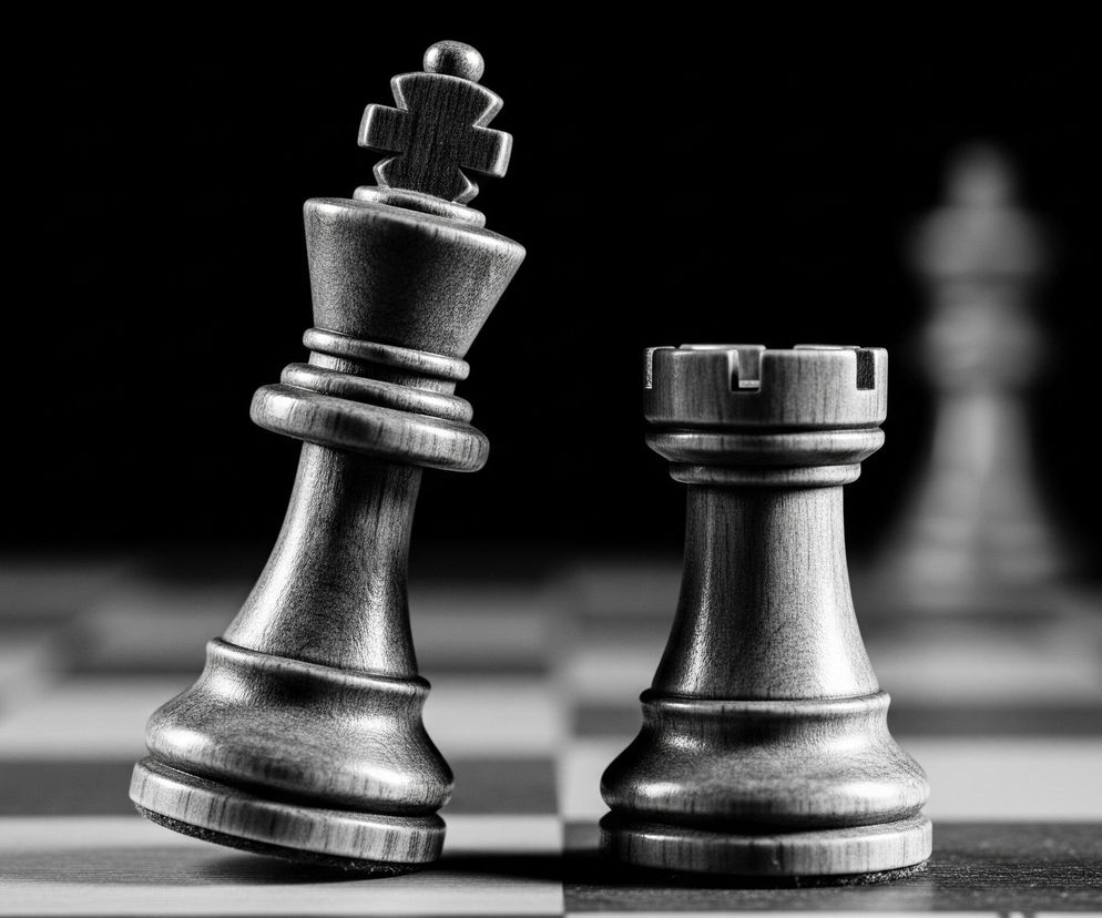 Dwie figury szachowe: króla i wieżę, ustawione na szachownicy w odcieniach szarości. Król, przechylony mocno w lewo, z wyraźną teksturą drewna, jest głównym obiektem w centrum kadru, oświetlonym od przodu. Obok niego stoi ciemna wieża, wyraźnie zarysowana i prosta. Tło jest czarne, rozmyte, z niewyraźnymi sylwetkami innych figur szachowych, zaś szachownica z białymi i czarnymi polami rozciąga się w dół, tworząc plan bliski.