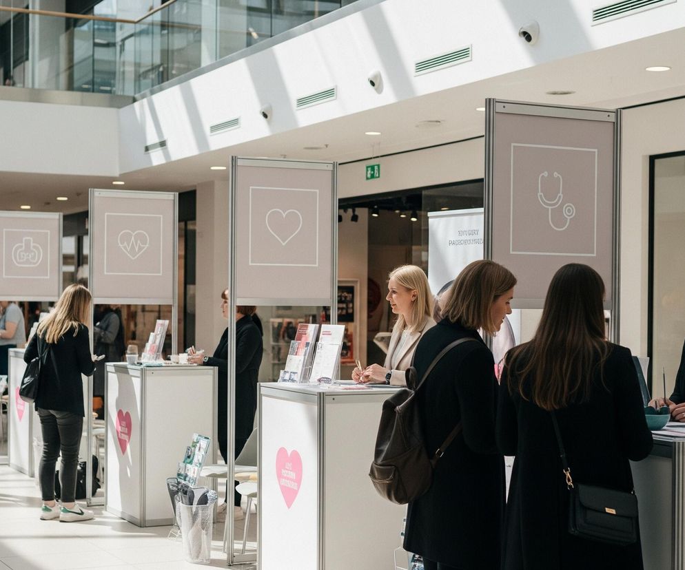 W przestronnym wnętrzu budynku, przypominającym pasaż handlowy, rozstawiono szereg białych stoisk z metalowymi ramami. Na każdym stoisku znajdują się pionowe panele w kolorze beżowym z białymi, stylizowanymi ikonami, takimi jak serce, linia EKG i stetoskop, a także białymi prostokątnymi ramkami. Przy stoiskach gromadzą się ludzie, głównie kobiety, niektóre z nich rozmawiają z obsługą, inne oglądają materiały promocyjne. Podłoga wyłożona jest jasnymi płytkami, przeplatanymi szarymi pasami, a w tle widać witryny sklepowe z odbijającymi się w nich światłami sufitowymi.