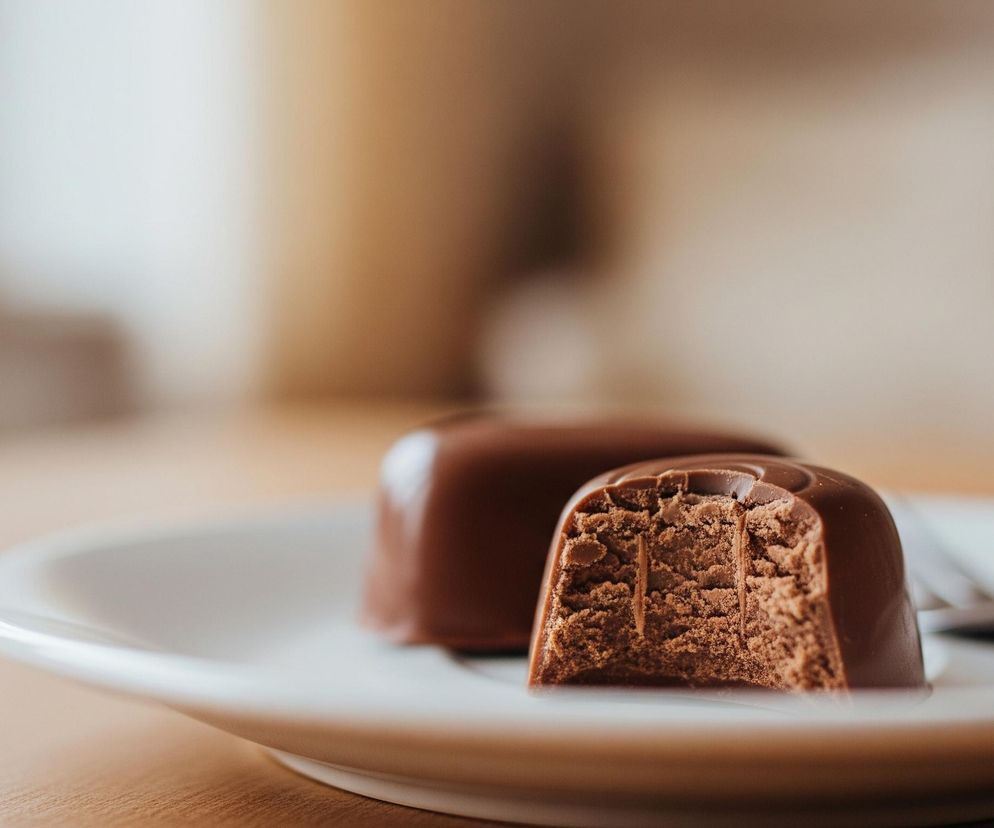 Na pierwszym planie, z prawej strony, leży na białym talerzyku czekoladowy cukierek, częściowo przełamany. Przełamany fragment ukazuje jasnobrązowe, ziarniste wnętrze z dwoma pionowymi, ciemniejszymi wgłębieniami. Za nim znajduje się drugi, nienaruszony cukierek o gładkiej, błyszczącej powierzchni. Na talerzyku, po prawej stronie za cukierkami, widać fragment srebrnego widelca. Talerzyk spoczywa na drewnianej powierzchni o ciepłym, beżowym odcieniu, rozjaśnionej po lewej stronie. Tło jest rozmyte, z dominującymi jasnymi, pastelowymi barwami, przechodzącymi od bieli po lewej do ciepłego brązu po prawej, z delikatnymi, ciemniejszymi akcentami.
