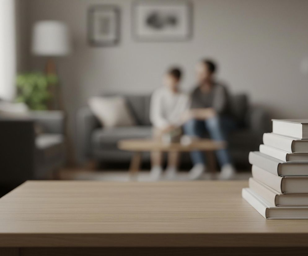 Na pierwszym planie, z prawej strony, na drewnianym blacie stołu, ułożony jest stos siedmiu książek o jasnych, niemal białych okładkach. Książki mają jednolitą grubość i są ułożone równo jedna na drugiej. Blat stołu jest w kolorze jasnego drewna z widoczną delikatną strukturą słojów.

Tło obrazu jest rozmyte, ukazując wnętrze pokoju. W centrum kadru, na szarej kanapie, siedzą dwie niewyraźne, ludzkie sylwetki, prawdopodobnie osoby dorosłe, zwrócone w lewo. Przed nimi, również rozmyty, widoczny jest mały, drewniany stolik kawowy. Po lewej stronie tła znajduje się okno z jasnymi zasłonami, a obok niego lampa stojąca z białym abażurem. Na ścianie za kanapą wiszą dwa rozmyte obrazy w ramkach. Całość wnętrza utrzymana jest w stonowanych, neutralnych barwach beżu, szarości i brązu.