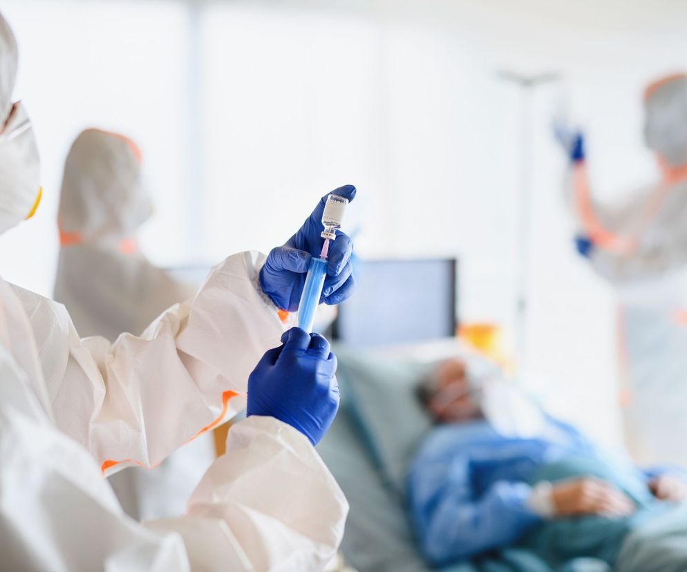 Osoba w kombinezonie ochronnym i niebieskich rękawicach przygotowuje szczepionkę w strzykawce, pobierając płyn z fiolki. W tle widać pacjenta w masce tlenowej i innego pracownika medycznego. Opracowywanie szczepionek to ważny temat, o którym więcej przeczytasz na Poradnik Zdrowie.