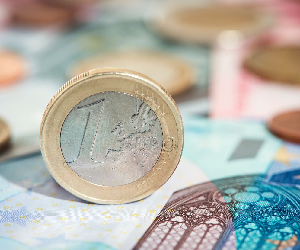 Moneta jednego euro stojąca pionowo na tle rozmazanych banknotów euro i innych monet. Zdjęcie symbolizuje unijne fundusze i rewizję KPO, o której możesz przeczytać na Super Biznes.