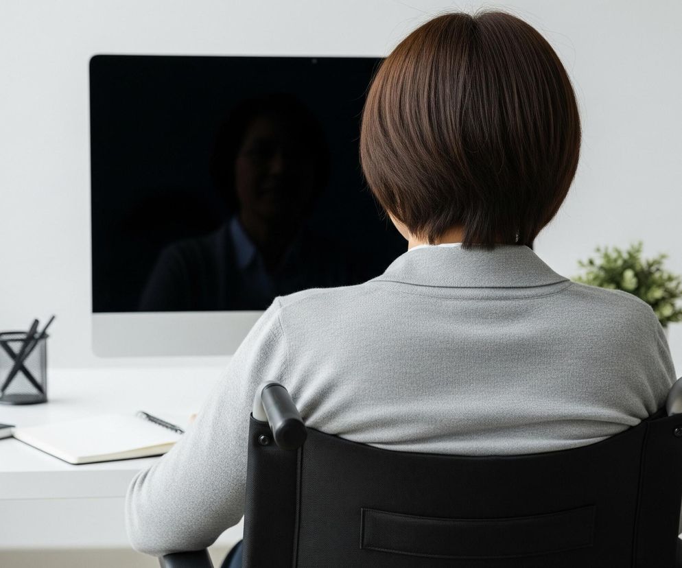 Osoba z krótkimi, ciemnymi włosami siedzi tyłem do widza w czarnym wózku inwalidzkim przed białym biurkiem. Na biurku, po lewej stronie, znajdują się dwa zeszyty, czarny pojemnik z trzema długopisami oraz otwarty notatnik z leżącym na nim długopisem. Centralnie umieszczony jest wyłączony monitor komputera z odbiciem postaci. Po prawej stronie biurka widnieje doniczka z zieloną rośliną oraz fragment białego regału z szarymi segregatorami.