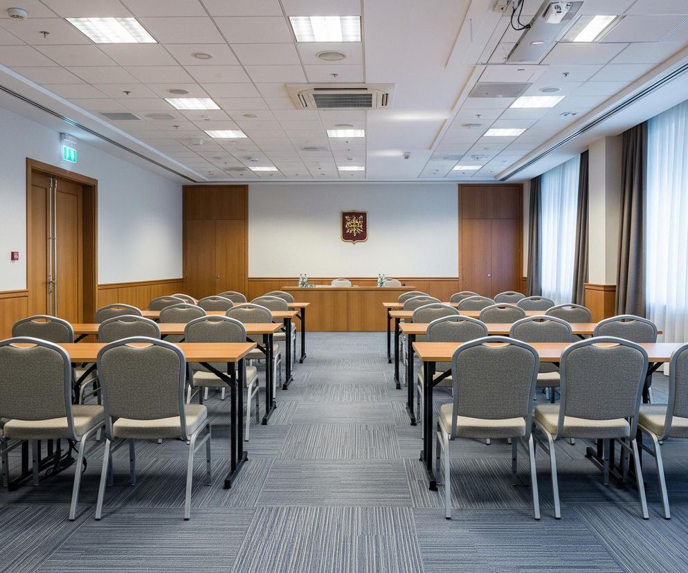 Wnętrze przestronnej sali konferencyjnej z ułożonymi rzędami stołów i krzeseł, przygotowanej do spotkania. Po obu stronach widoczne są dwa rzędy podłużnych, jasnobrązowych stołów, przy których stoją szare krzesła z metalowymi stelażami, z czego w każdym rzędzie jest po dziewięć stołów i krzesła po obu ich stronach. Ściany boczne mają dolną część boazerią w kolorze drewna, a górną białą, natomiast ściana główna, na której wisi czerwono-złoty herb, jest cała boazerią. Na suficie podwieszanym, podzielonym na kwadratowe panele, zamontowano liczne prostokątne i okrągłe oprawy oświetleniowe, a także kratki wentylacyjne, z centralnie umiejscowionym kwadratowym otworem klimatyzacji. Po prawej stronie widać duże okno zakryte jasnymi, półprzezroczystymi firankami oraz zasłonami w kolorze ciemnoszarym, a po lewej stronie znajdują się podwójne, drewniane drzwi, nad którymi wisi podświetlony zielony znak wyjścia ewakuacyjnego. Na końcu sali, na podniesieniu, stoi stół prezydialny z dwoma butelkami wody i szklankami, za którym znajduje się wspomniany herb. Podłoga pokryta jest dywanową wykładziną w odcieniach szarości i błękitu, z drobnymi, równoległymi paskami, ułożoną w naprzemienne kwadraty.