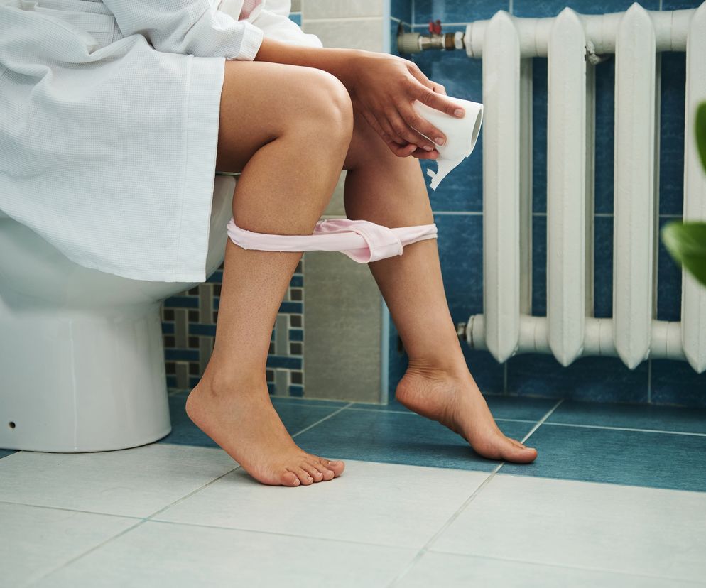 Zbliżenie na kobietę w łazience, która siedzi na WC, w ręce trzyma rolkę papieru toaletowego. 