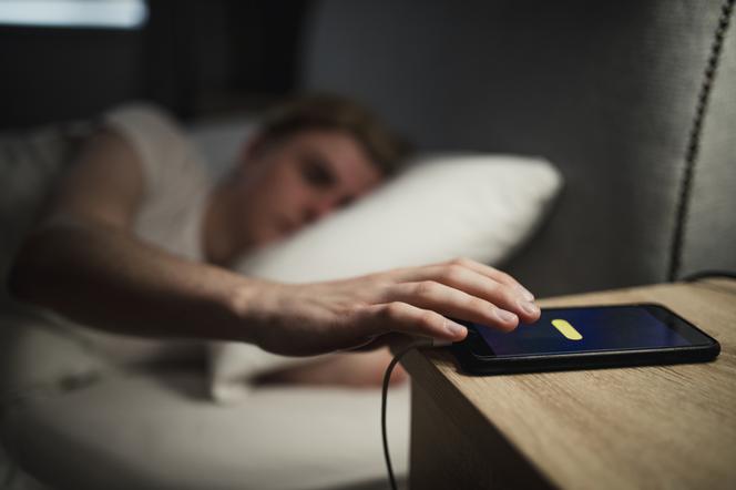 mężczyzna leżący na łóżku sięga po telefon znajdujący się na szafce nocnej