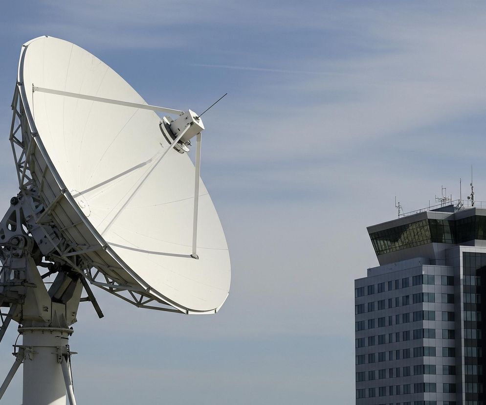 Po lewej stronie kadru znajduje się duża, biała antena satelitarna, składająca się z okrągłej, parabolicznej czaszy i stalowej konstrukcji wsporczej z drabiną. Po prawej stronie widać wysoki, nowoczesny budynek w odcieniach szarości i bieli, z licznymi rzędami ciemnych okien i przeszkloną, pochyloną konstrukcją na dachu. Oba obiekty są ukazane na tle jasnoniebieskiego nieba z delikatnymi smugami chmur.