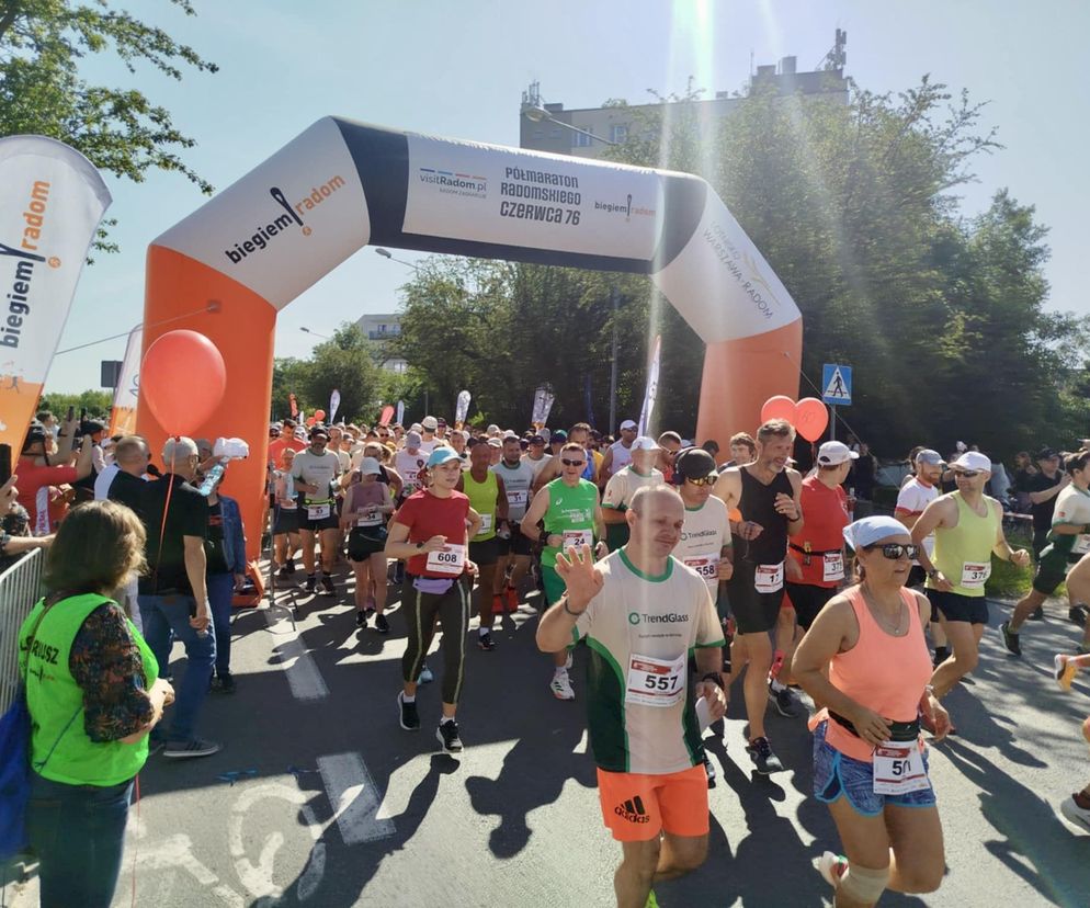 Uczestnicy Półmaratonu Radomskiego Czerwca '76 biegnący pod pomarańczową bramą startową z napisem biegiem radom, z numerami startowymi, podczas słonecznego dnia. Tę relację z wydarzenia przeczytasz na naszym portalu.