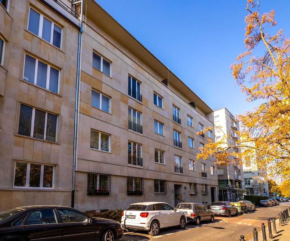 Modernistyczna kamienica przy ul. Konopnickiej 5 w Warszawie po remoncie, z elewacją z piaskowca, licznymi oknami i balkonami z metaloplastyką. Widoczne zaparkowane samochody oraz drzewa z jesiennymi liśćmi, symbolizujące luksus i historię obiektu, o czym można przeczytać na Architektura Murator Plus.