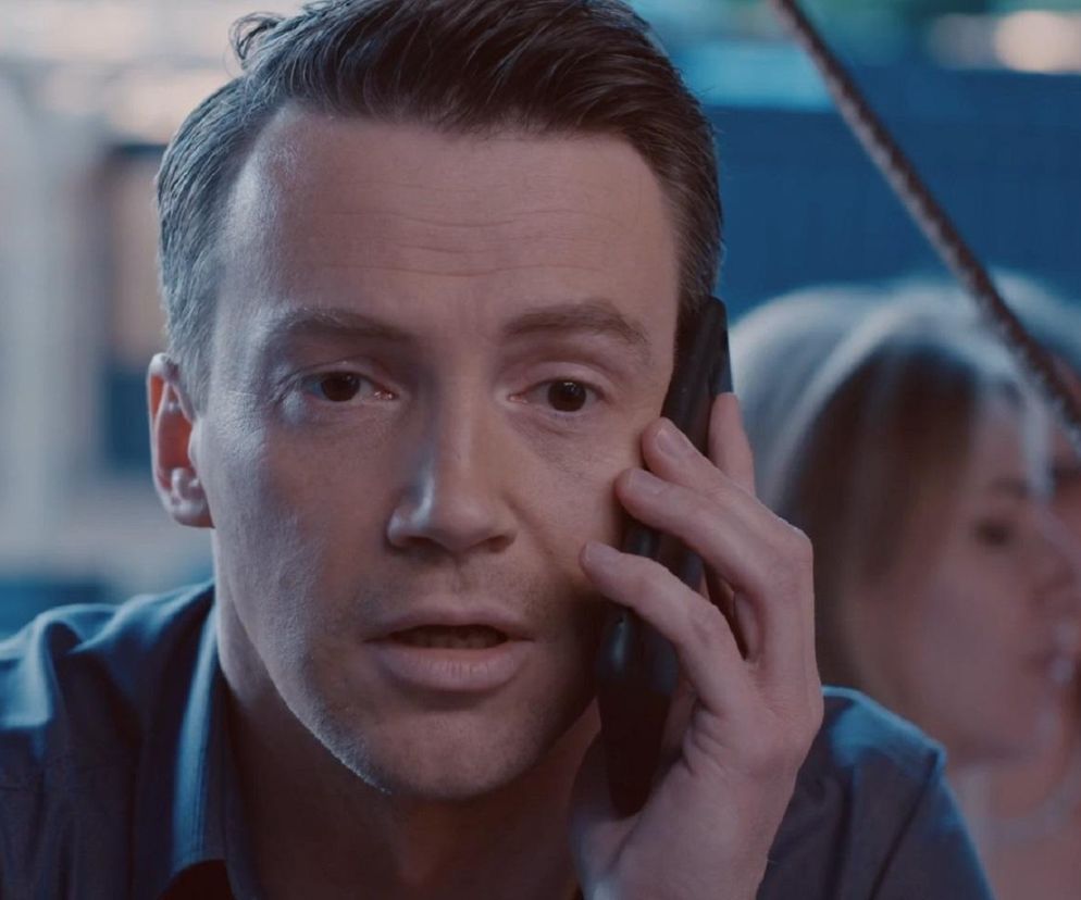 Aktor Krzysztof Kwiatkowski jako Jakub z serialu M jak miłość podczas rozmowy telefonicznej, w tle widać rozmazaną postać innej osoby. Scena ukazuje jego załamanie na wieczorze kawalerskim Marcina, co podkreśla dramatyzm wydarzeń. Więcej o losach bohatera przeczytasz na stronie Super Seriale.