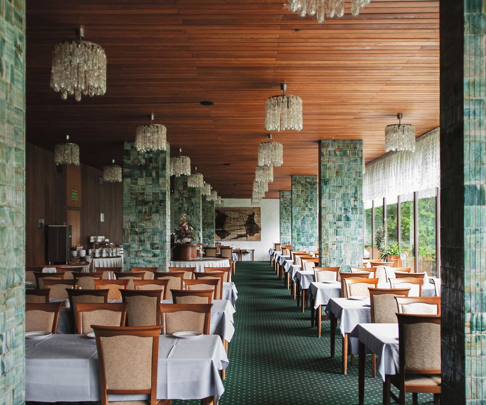 Wnętrze restauracji z połowy XX wieku, charakteryzujące się długimi rzędami stołów z białymi obrusami i drewnianymi krzesłami, zielonym wzorzystym dywanem oraz drewnianym sufitem z licznymi szklanymi żyrandolami. Ściany i kolumny pokryte są turkusowymi płytkami, a w tle widać duże okna z widokiem na zieleń. Takie obiekty i ich architekturę z czasów PRL omawia Architektura Murator Plus.