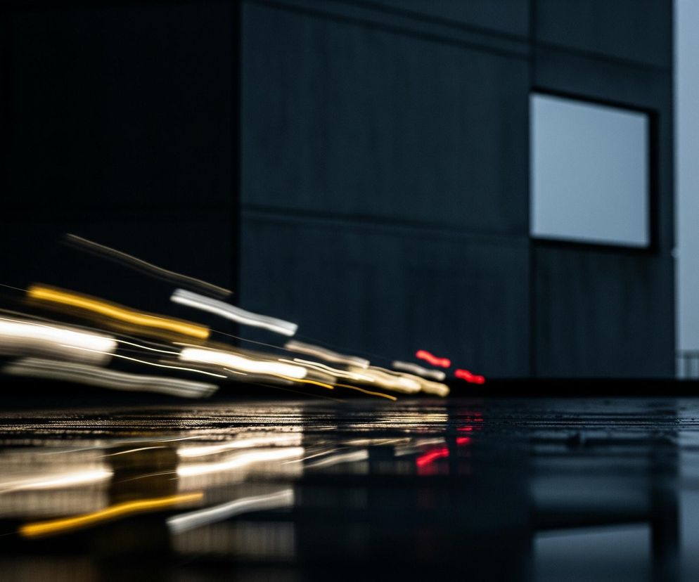 Na pierwszym planie widoczne są rozmyte smugi światła o barwie białej, żółtej i czerwonej, ciągnące się ukośnie od lewej do prawej strony, znikając w tle. Poniżej smug znajduje się ciemna, lśniąca powierzchnia, odbijająca te światła, co sugeruje wilgotną nawierzchnię. W tle po lewej stronie dominuje ciemna, gładka ściana budynku, a po prawej stronie tej ściany znajduje się pojedyncze, prostokątne okno o jasnej, lekko zamglonej szybie. Górna prawa część obrazu jest zdominowana przez jasnoszare, jednolite niebo, tworzące tło dla horyzontu.