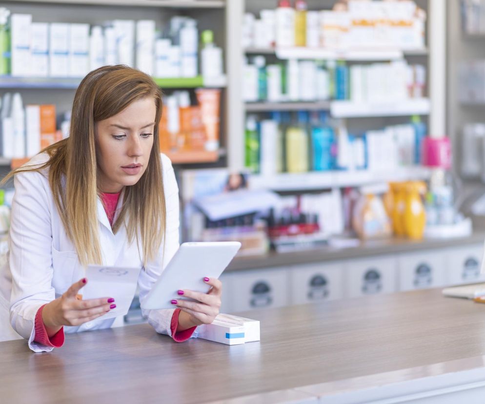 Kobieta w białym fartuchu farmaceutki z tabletem i receptą w rękach, stojąca za ladą apteczną, z półkami pełnymi leków w tle. Ilustruje temat braków leków na rynku, o których więcej przeczytasz na Poradnik Zdrowie.