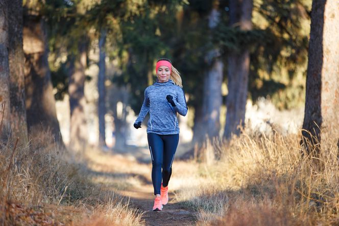 Kobieta w sportowym stroju, biegnąca leśną ścieżką. Ma na sobie niebieską bluzę z kapturem, czarne legginsy, różową opaskę na głowie i różowe buty, co doskonale ilustruje aktywność fizyczną. Artykuł na Poradnik Zdrowie dotyczy wyboru bezprzewodowych słuchawek do biegania.