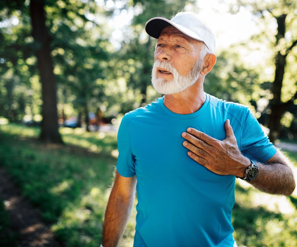 Starszy mężczyzna z białą brodą i wąsami, w niebieskiej koszulce i białym daszku, trzymający dłoń na klatce piersiowej, stojący w parku. Wygląda na zmęczonego po wysiłku fizycznym. Dowiedz się, jak zachować formę po 60-tce na Poradnik Zdrowie.