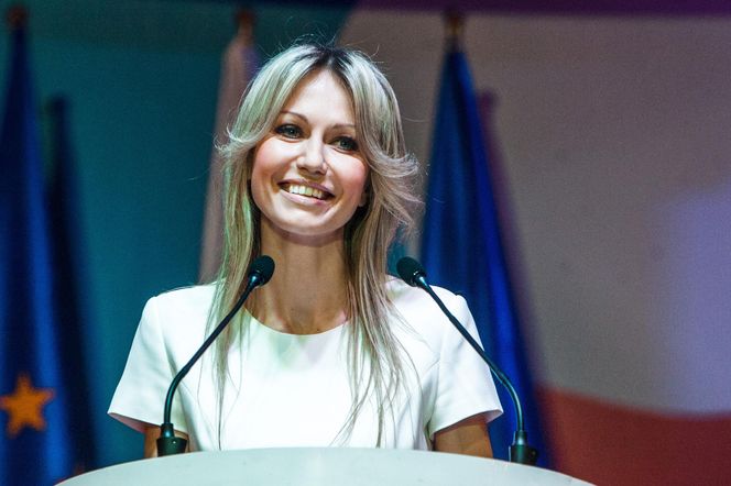 Magdalena Ogórek, prezenterka, uśmiecha się podczas wystąpienia publicznego, stojąc za mównicą z mikrofonami, z europejskimi flagami w tle. Artykuł na Poradnik Zdrowie dotyczy jej opieki nad chorym byłym mężem.