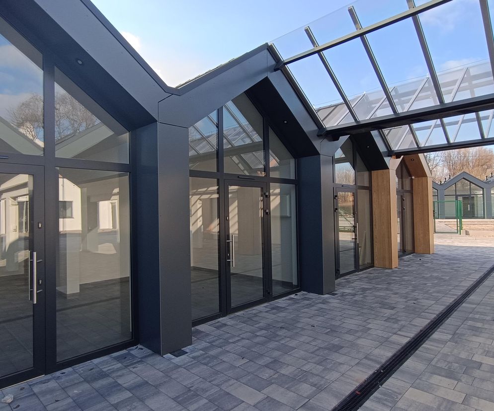 Nowoczesne pawilony handlowe z przeszklonymi frontami i szarymi, spadzistymi dachami tworzące część kompleksu targowiska miejskiego w Brześciu Kujawskim. Na pierwszym planie widoczne są szare płytki chodnikowe. Więcej o architekturze tego typu przeczytasz na Architektura Murator Plus.