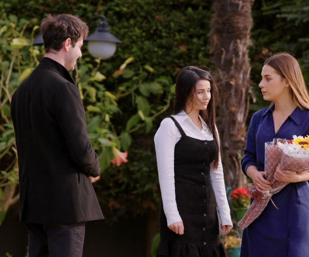 Młody mężczyzna w czarnej marynarce, zwrócony tyłem, rozmawia z dwiema kobietami w ogrodzie. Kobieta po lewej w białej koszuli i czarnej sukience spogląda w dół, a kobieta po prawej w niebieskiej sukience trzyma bukiet białych kwiatów i słonecznika. Możesz przeczytać o losach bohaterów serialu Miłość i nadzieja na portalu Super Seriale.
