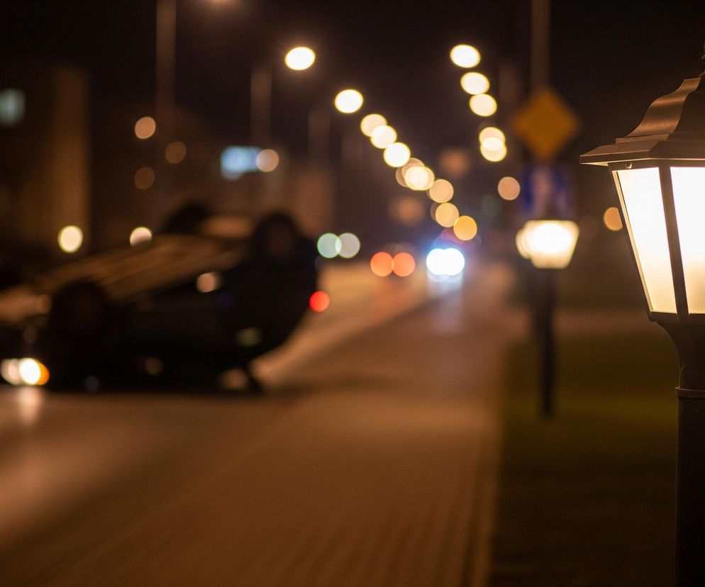 Na pierwszym planie, po prawej stronie, widać oświetloną latarnię uliczną w stylu retro, z metalowym czarnym korpusem i mlecznymi szybami. Światło z latarni intensywnie oświetla chodnik i część trawnika po prawej stronie kadru. W tle, na rozmytej ulicy, leży na boku przewrócony samochód, a jego światła awaryjne są widoczne jako jasne punkty. Dalsze tło wypełniają liczne rozmyte światła innych latarni ulicznych oraz świateł pojazdów, tworząc efekt bokeh, z dominującymi odcieniami żółci, pomarańczu i czerwieni.