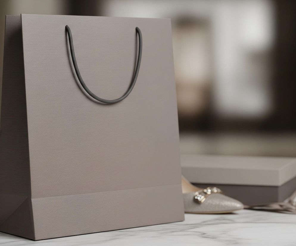 Na pierwszym planie, centralnie, stoi duża, prostokątna torba na zakupy w odcieniu szaro-brązowym, z matową, lekko teksturowaną powierzchnią i ciemniejszymi sznurkowymi uchwytami. Po lewej stronie torby, na szarym pudełku z pokrywką, leży lśniący szampański szpilkowy but ze złotymi i przezroczystymi ozdobami na nosku. Po prawej stronie, w tle, widoczny jest fragment innego szampańskiego buta, leżącego obok otwartego szarego pudełka z pokrywką i luźno rozłożonej wstążki w dopasowanym kolorze. Całość kompozycji spoczywa na białym, marmurowym blacie z delikatnymi szarymi żyłkami, a tło jest rozmyte, ukazując niewyraźne, jasne i ciemne kształty.