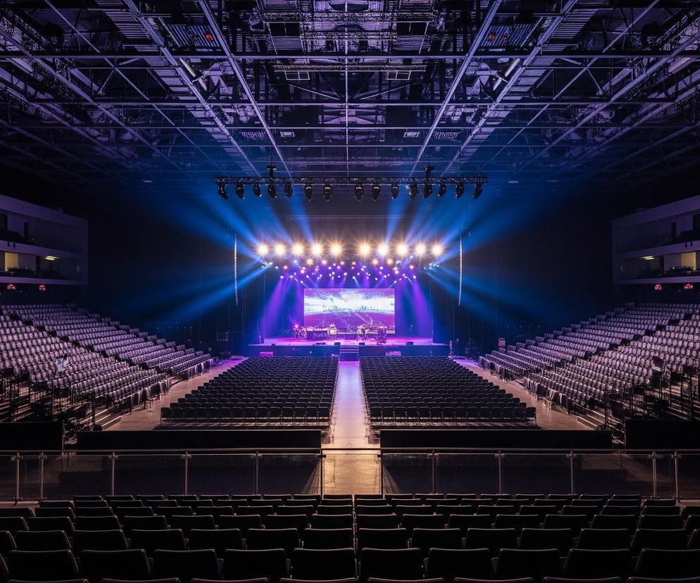 Pusta sala koncertowa z rzędami czarnych siedzeń rozciągających się po obu stronach centralnej sceny. Scena jest jasno oświetlona fioletowymi i niebieskimi światłami, tworząc wyraźne smugi światła, a na jej tle widoczny jest duży ekran wyświetlający abstrakcyjny wzór. Sufit sali jest odkrytą konstrukcją z kratownic i instalacji technicznych, oświetloną subtelnym, fioletowym światłem, kontrastującym z ciemnymi barwami widowni.