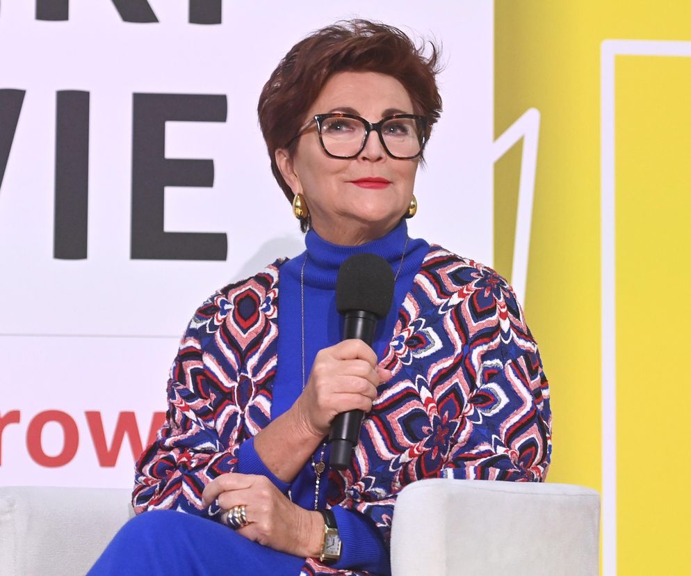Jolanta Kwaśniewska w eleganckich okularach w czarnych oprawkach i niebieskim golfie, narzucając na siebie wzorzystą narzutkę w odcieniach czerwieni, bieli i granatu. Prezentuje nowoczesny styl, o którym więcej przeczytasz na naszym portalu.