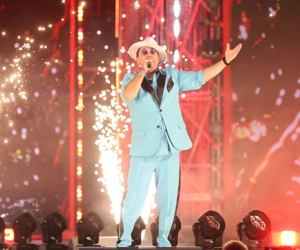 Marcin Miller, lider zespołu Boys, w błękitnym garniturze i kapeluszu, śpiewa na scenie, unosząc ręce. W tle widać efekty świetlne i iskry. Artykuł na Poradnik Zdrowie opisuje jego walkę z bólem kręgosłupa i codzienne wyzwania.