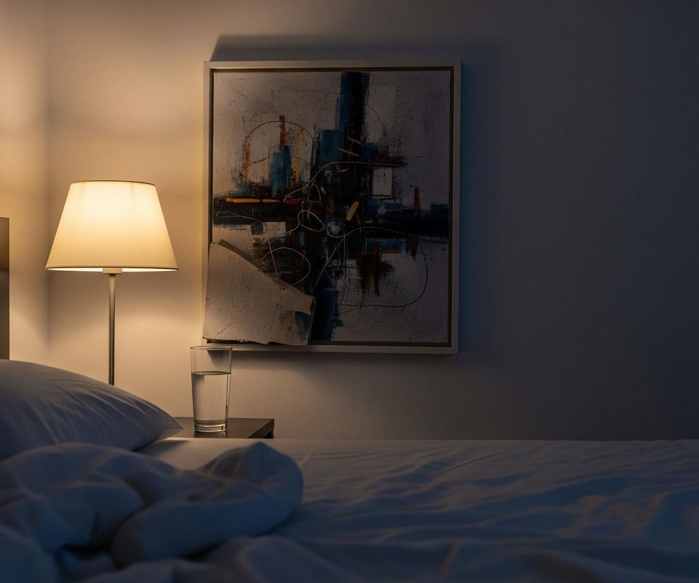 Scena przedstawia fragment sypialni o zmierzchu lub w nocy, z dominującymi odcieniami błękitu i szarości. Po lewej stronie widać kawałek ciemnego zagłówka łóżka, a na nim dwie poduszki oraz pogniecioną, białą pościel. Obok łóżka stoi nocna szafka, na której znajduje się szklanka z wodą oraz świecąca lampa stołowa z kremowym abażurem, rzucająca ciepłe, żółte światło na ścianę. Nad szafką, na ścianie, wisi prostokątny obraz w jasnej ramie, przedstawiający abstrakcyjną kompozycję z elementami niebieskimi, czarnymi, rdzawymi i białymi.