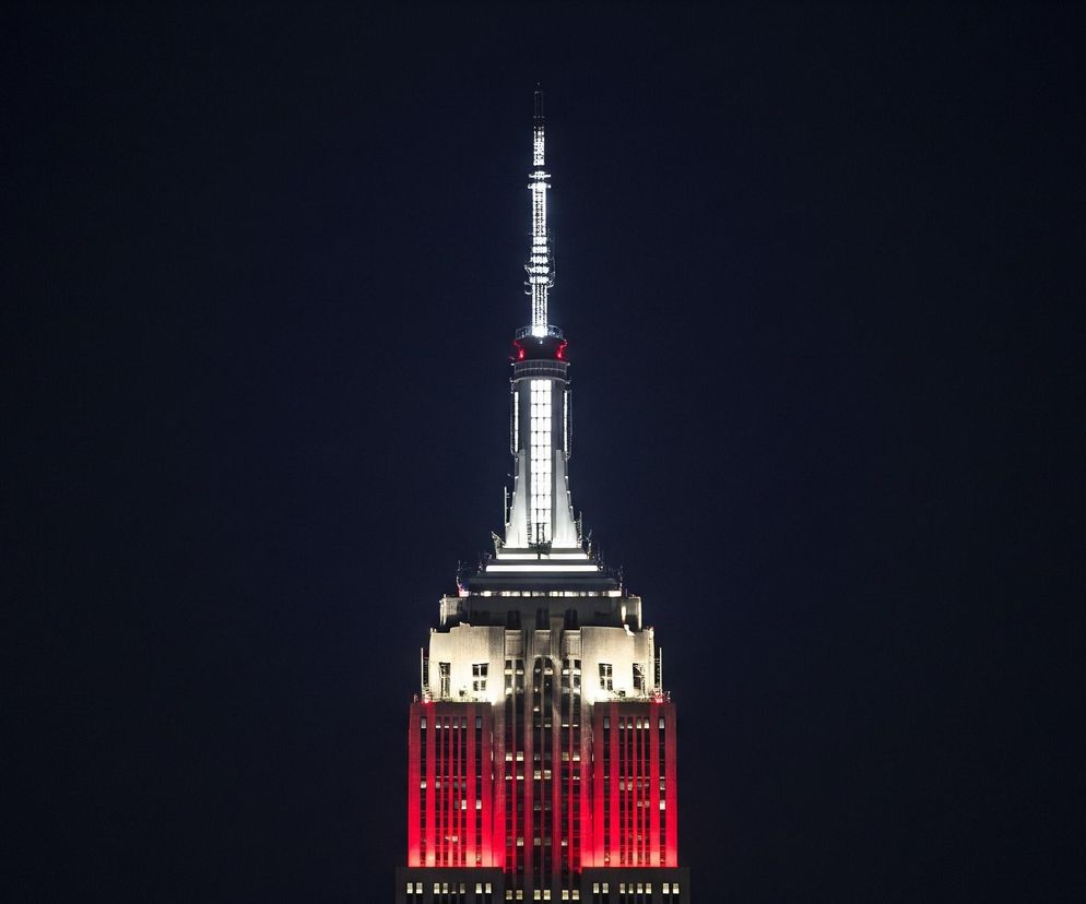 Strzelisty budynek zajmuje centralną część kadru, rozciągając się od dołu do góry na ciemnym, granatowym tle nocnego nieba. Górna część konstrukcji jest biała, z widocznymi poziomymi segmentami i pionową iglicą na szczycie, ozdobioną drobnymi, białymi światełkami. Poniżej, w środkowej części budynku, jasnoczerwone oświetlenie tworzy wyraziste pionowe pasy, kontrastujące z bielą górnych partii i niewielką, szarą sekcją poniżej. U samego dołu obrazu widoczne są pojedyncze, żółte punkty światła, sugerujące okna w niższych, ciemniejszych partiach konstrukcji.